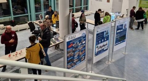 The photo shows a group of participants interacting with institute members, as well as 3 different posters describing research.